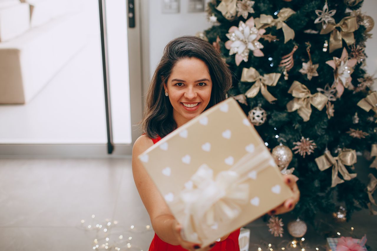 heros-img Smiling woman holding a Christmas gift in front of a decorated tree indoors.