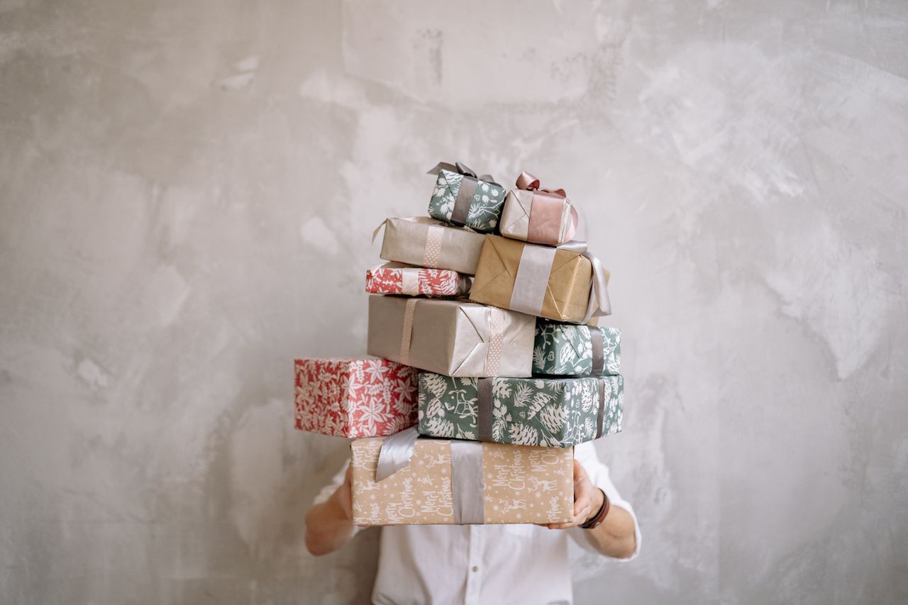 services-03 Person hiding behind a tall stack of gift-wrapped boxes against a gray wall, indoors.