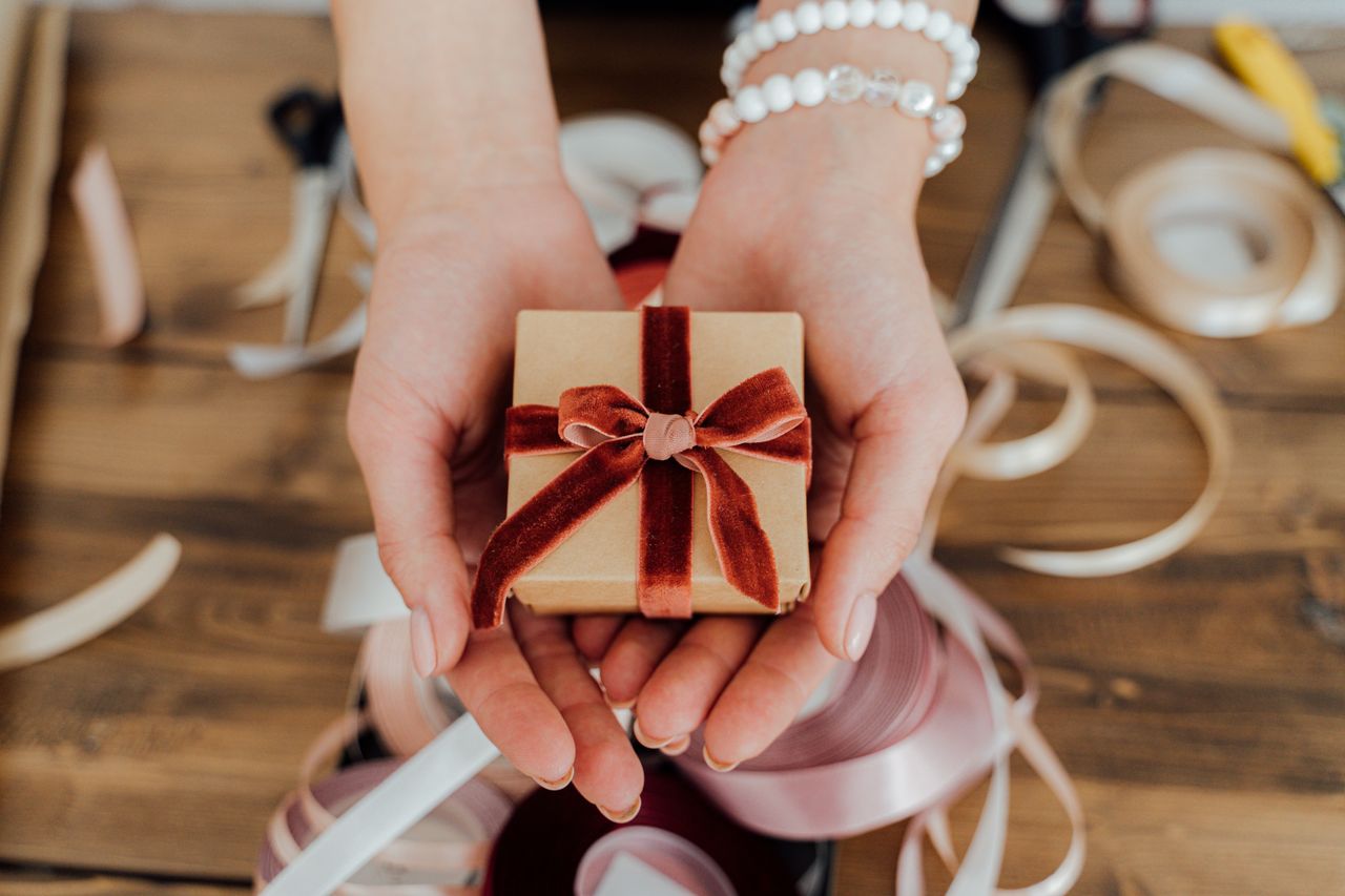 about-us Hands presenting a small gift box wrapped with red velvet ribbon, surrounded by crafting materials.