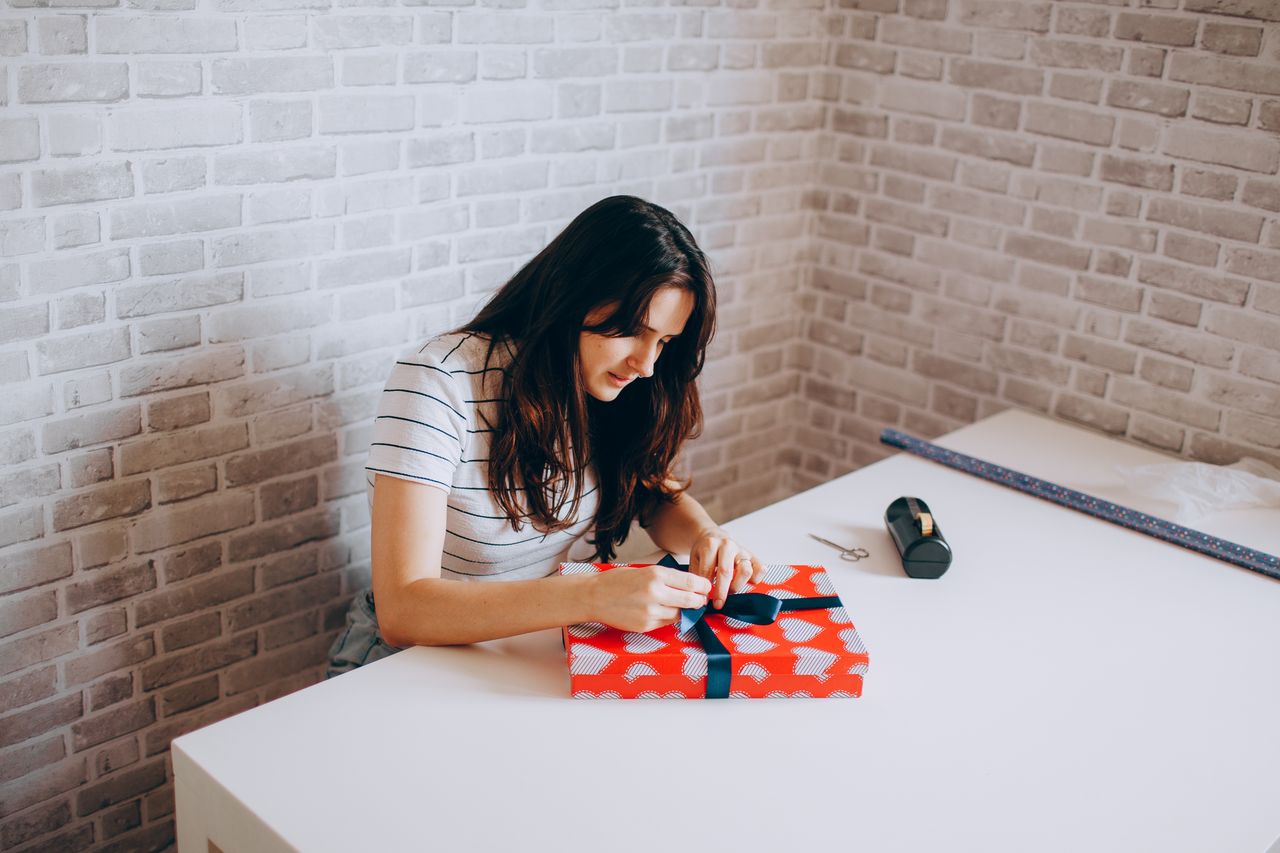 who-we-are Woman gift wrapping at table in cozy room with brick walls, showcasing creativity and leisure.